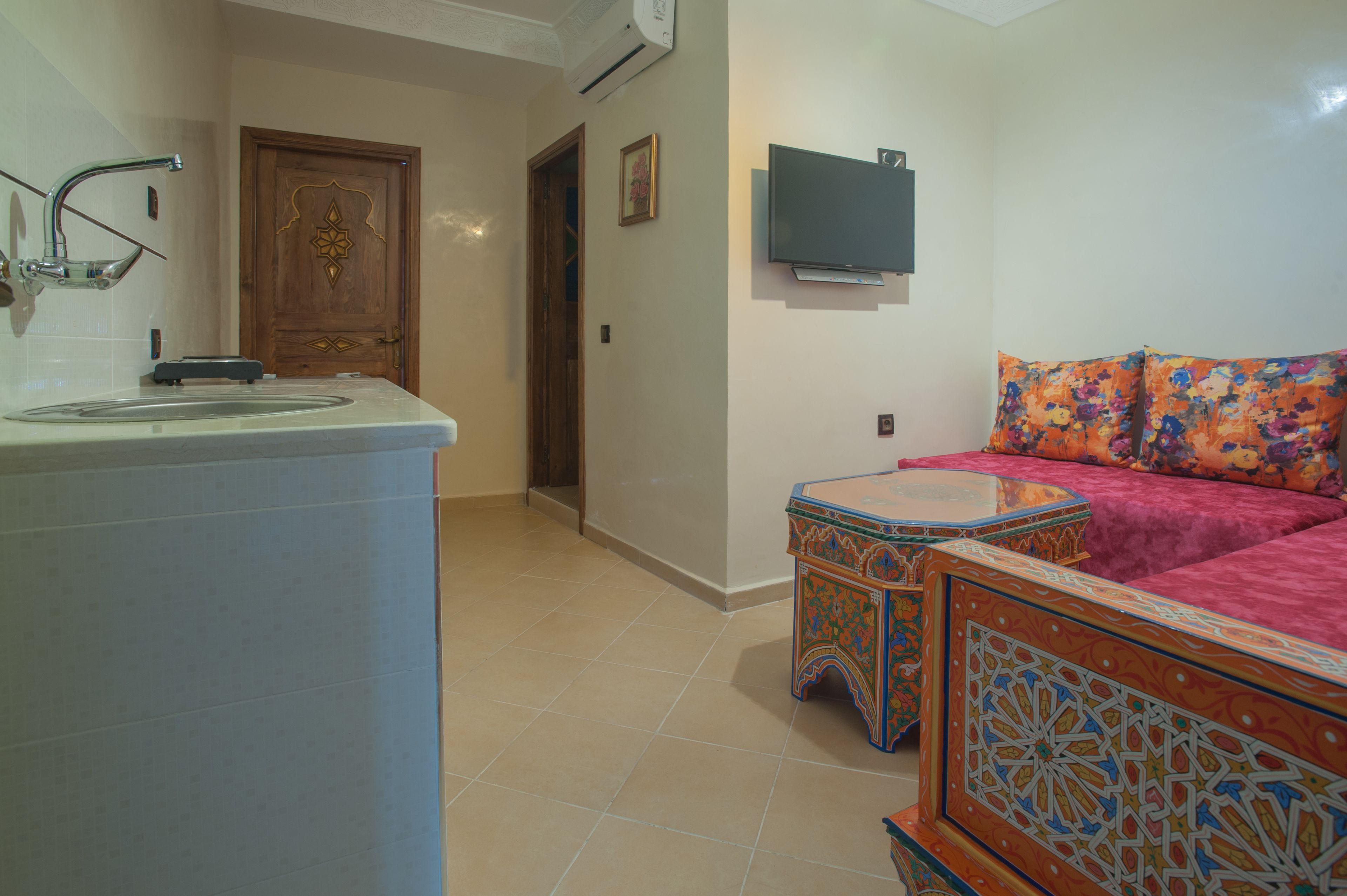 A room with a kitchen area, a wooden door, a TV mounted on the wall, and a sofa with floral cushions.