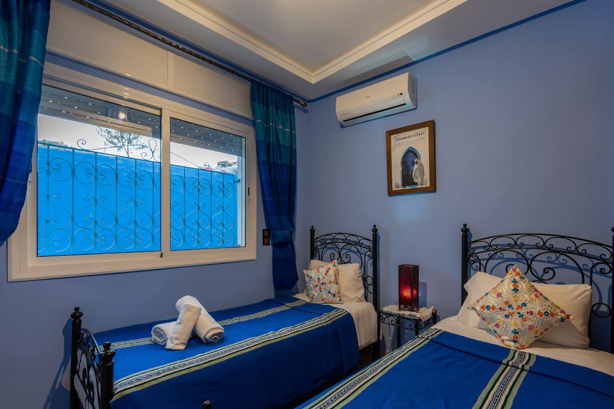 Un dormitorio con dos camas, una ventana con cortinas azules, un aire acondicionado y un cuadro en la pared.
