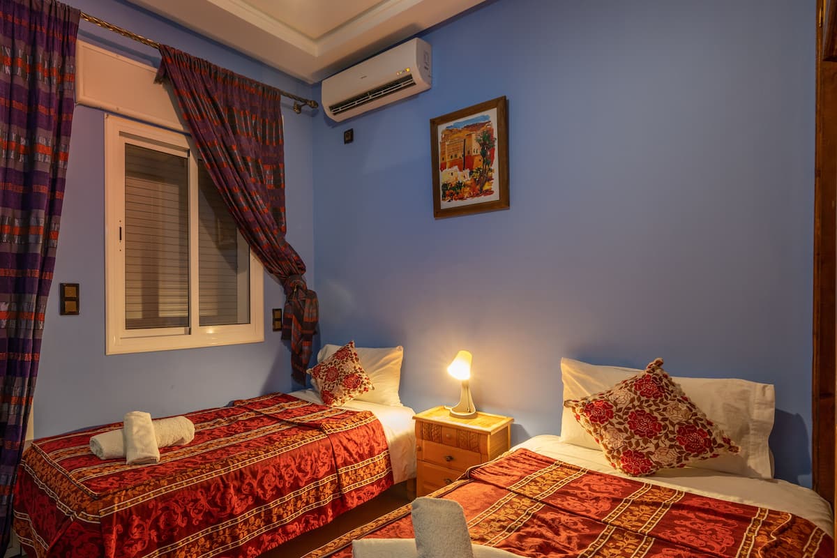 Una habitación con dos camas, cada una con ropa de cama estampada en rojo y almohadas blancas. Hay una ventana con cortinas, un equipo de aire acondicionado en la pared y una mesita con una lámpara.