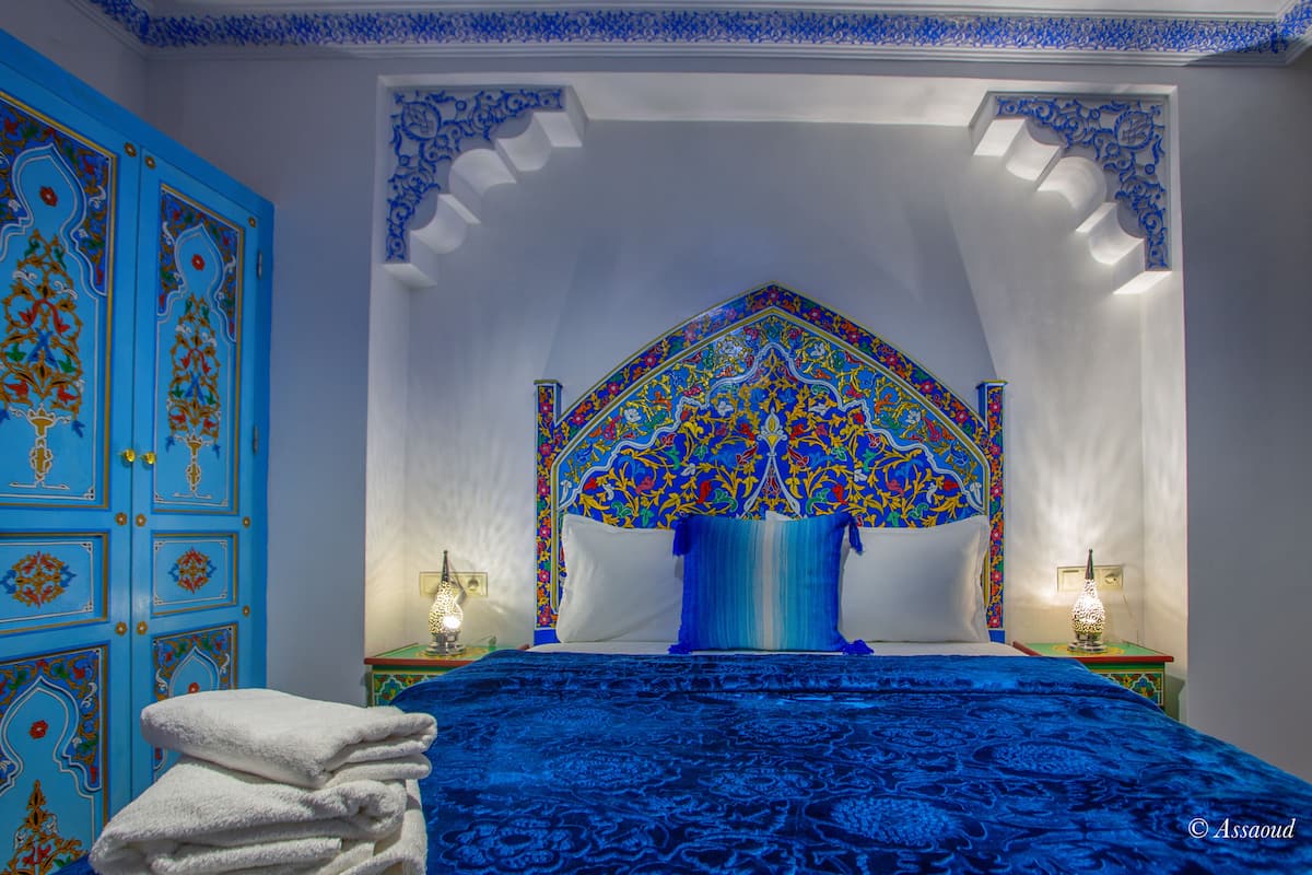 Una habitación con una cama, una puerta azul con diseños intrincados y un techo decorativo.