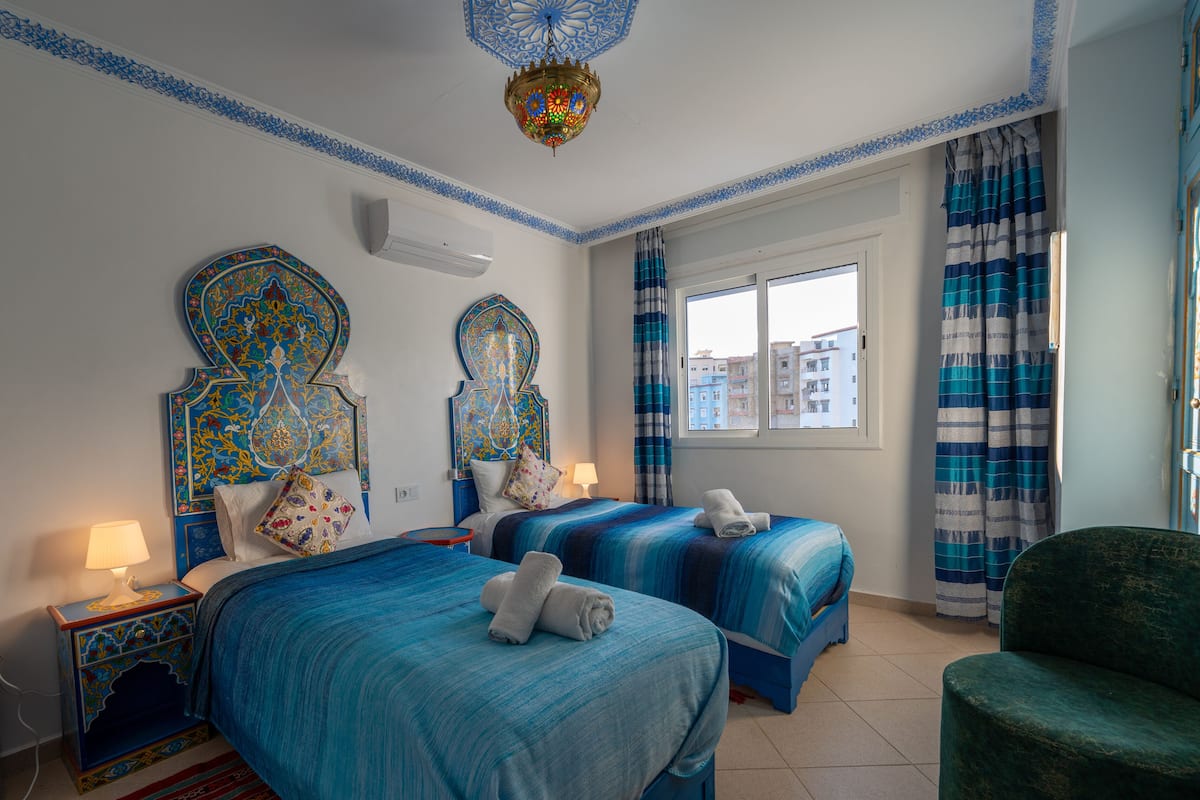 Habitación de hotel con dos camas, una lámpara decorativa en el techo, una ventana con cortinas y un sillón verde.
