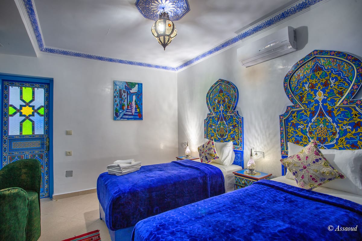 Una habitación con dos camas, una silla verde, un muro decorativo y una lámpara en el techo.