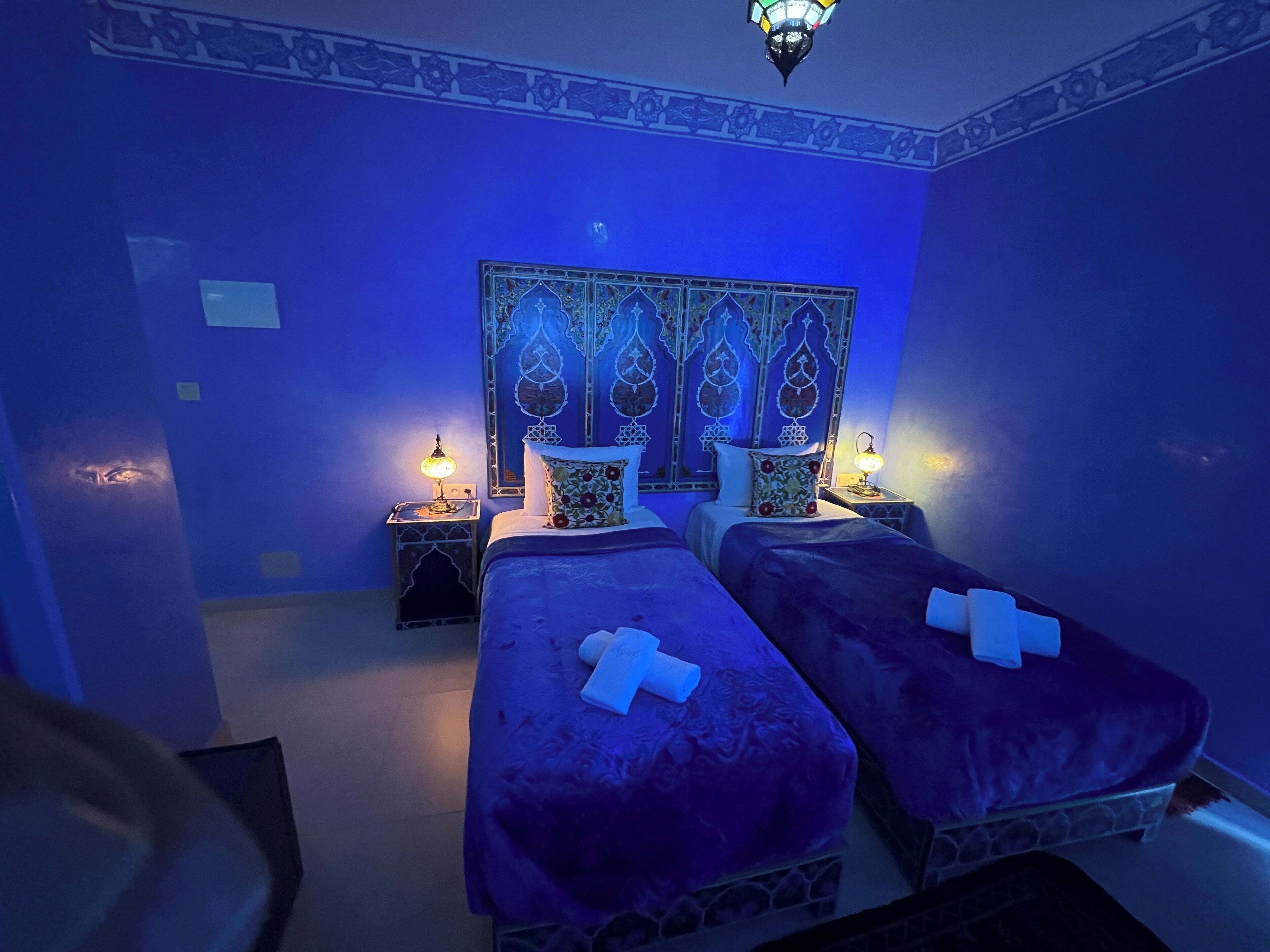 Una habitación con dos camas, cada una con cubrecama morado y una cruz blanca en las almohadas. Hay dos mesitas de noche con lámparas y un tapiz decorativo sobre las camas.