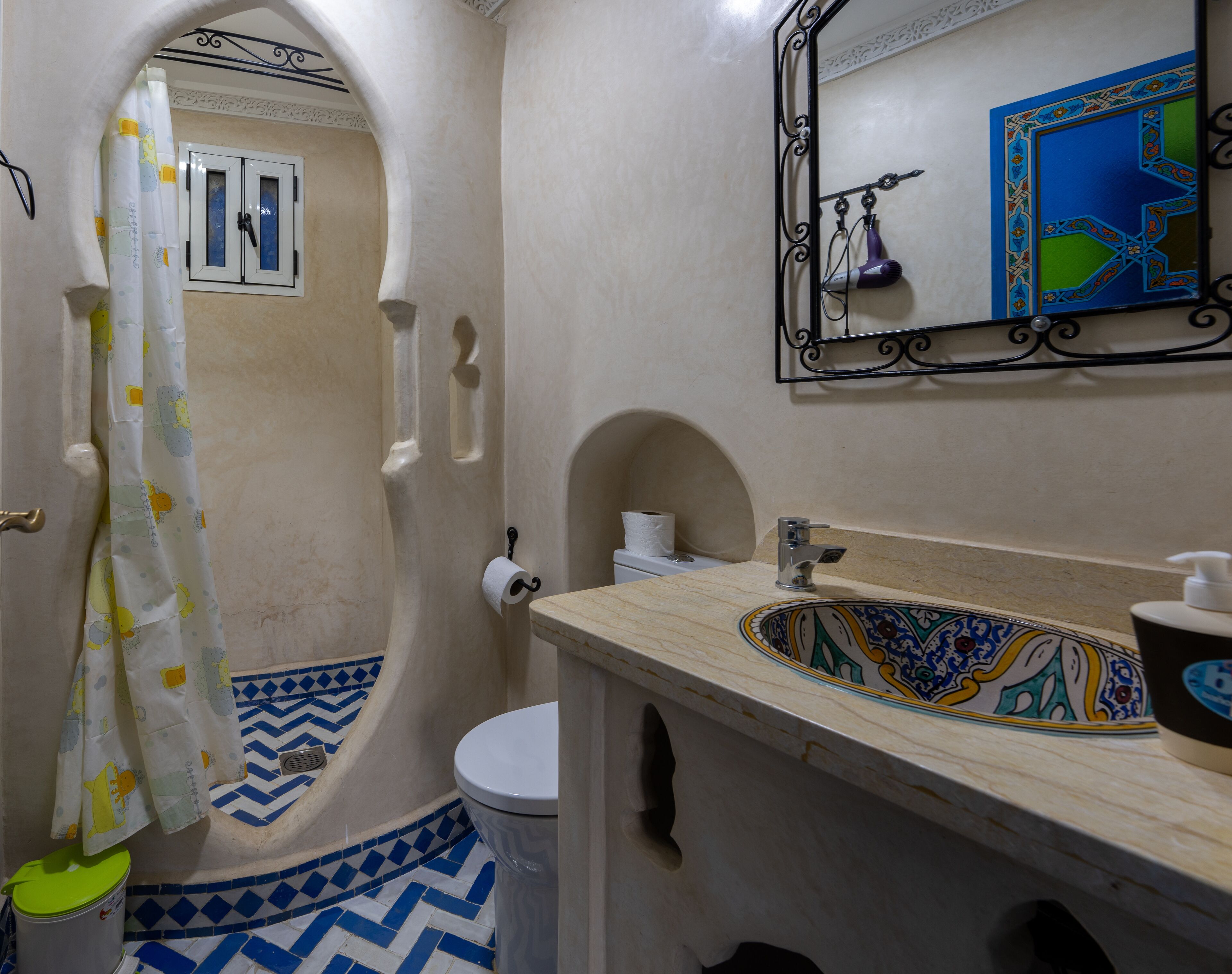 A bathroom with a white toilet, a sink with a decorative tile backsplash, a shower with a blue and white tile floor, and a mirror with a decorative frame.