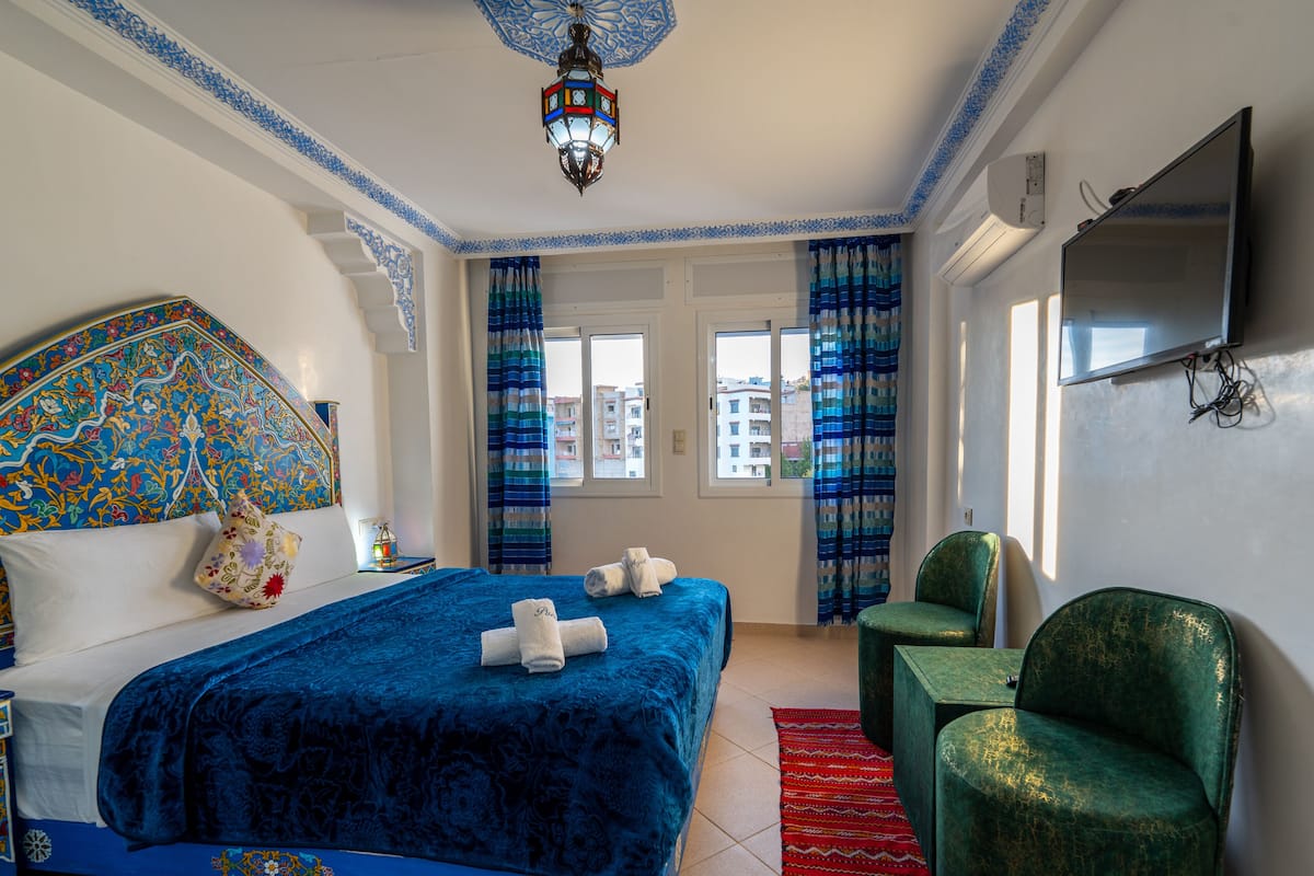 Una habitación de hotel con una cama, un sillón verde, un televisor y una lámpara decorativa en el techo.