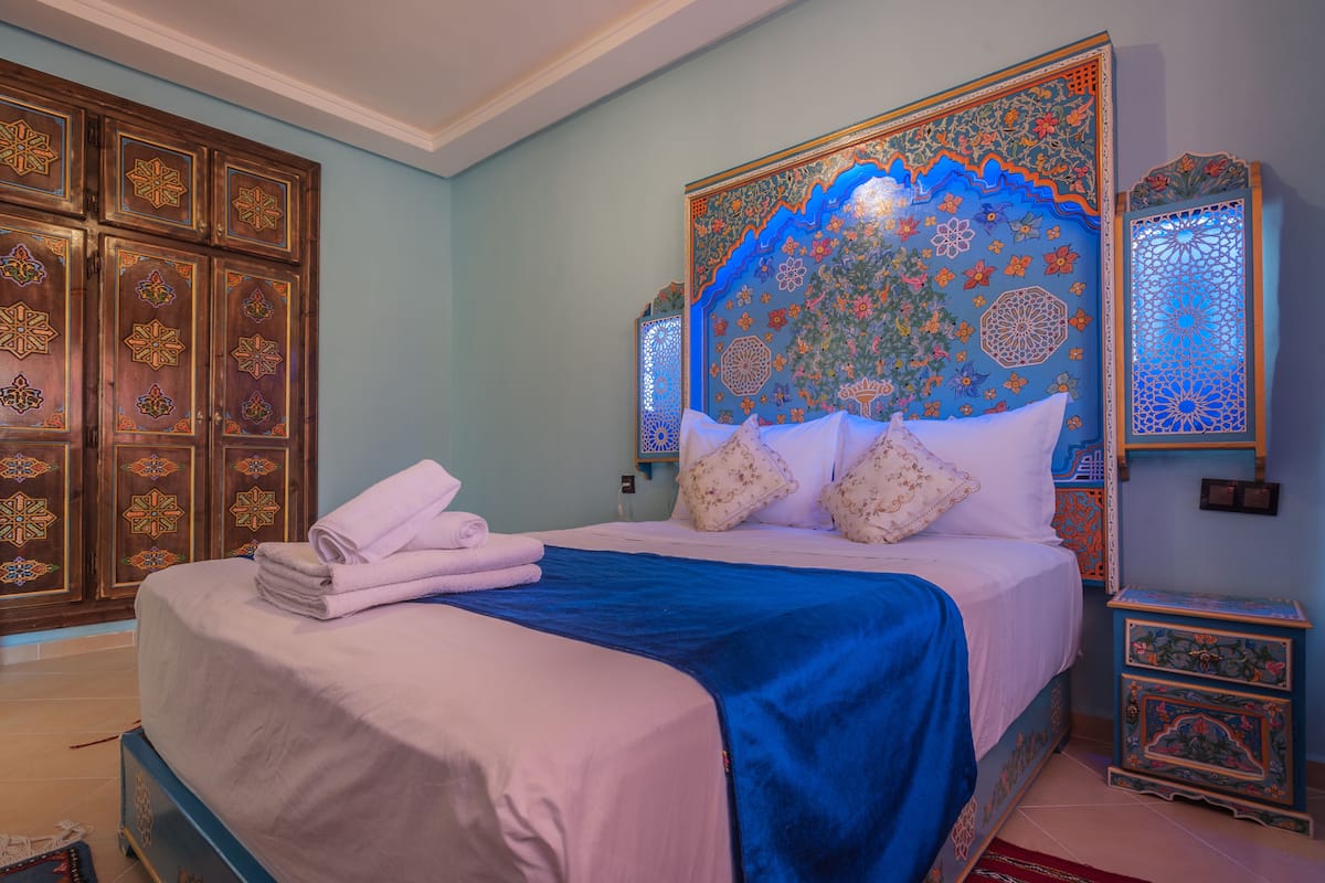 Una cama bien hecha con una colcha azul y almohadas blancas, un cabecero decorativo con diseños intrincados y un par de armarios de madera con detalles elaborados.
