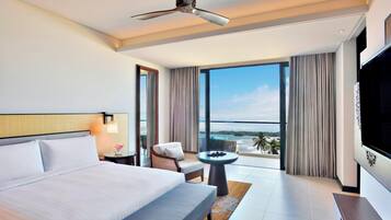 Habitación de hotel moderna con una cama grande, ventilador de techo, televisión, balcón con vista al océano y una zona de estar con una mesa.