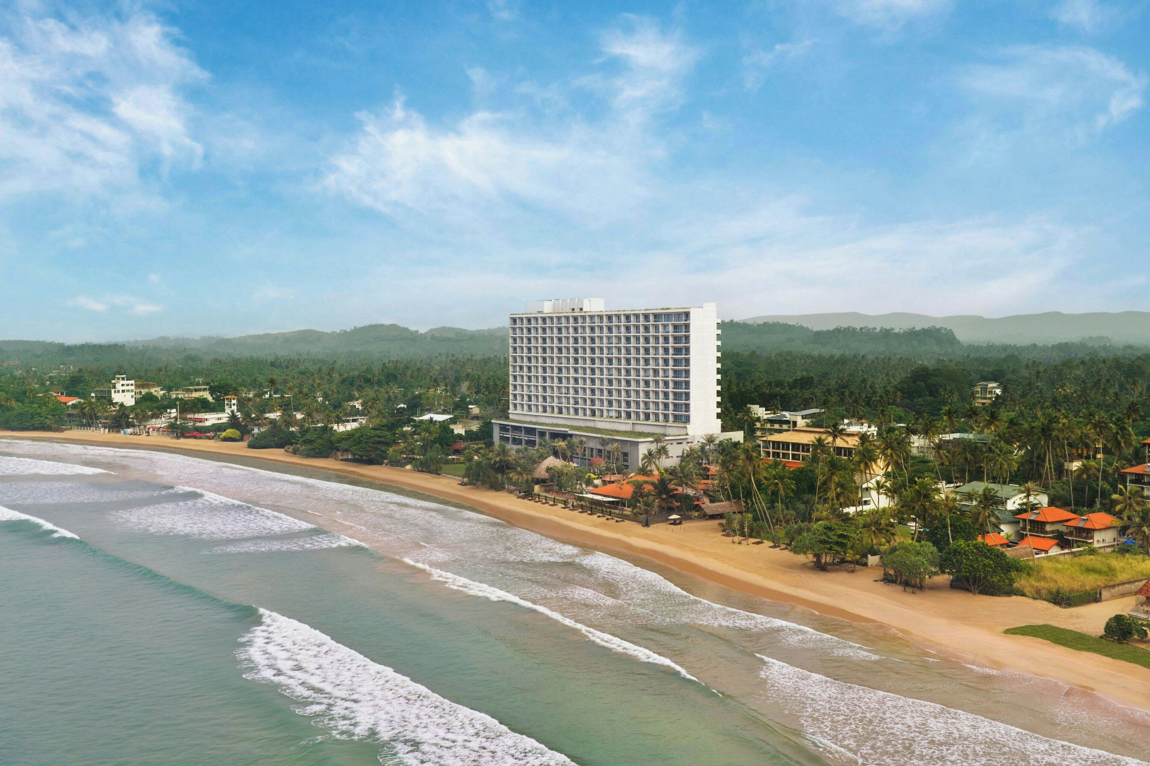 A beachfront hotel with a large building, a sandy beach, and a forested background.