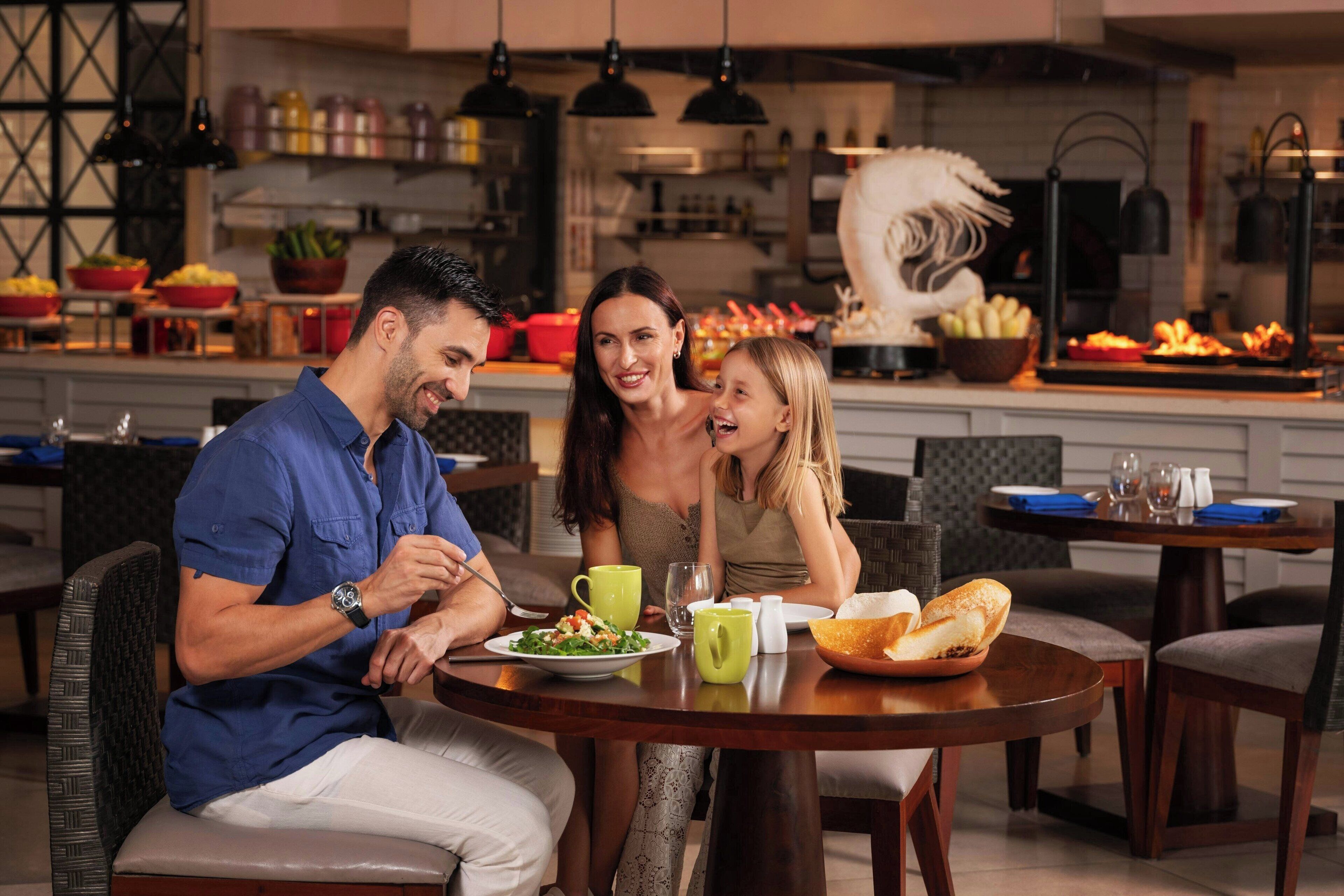 Una familia comiendo en una mesa redonda en una cocina moderna con una isla central, sillas de comedor y una variedad de utensilios y electrodomésticos de cocina al fondo.