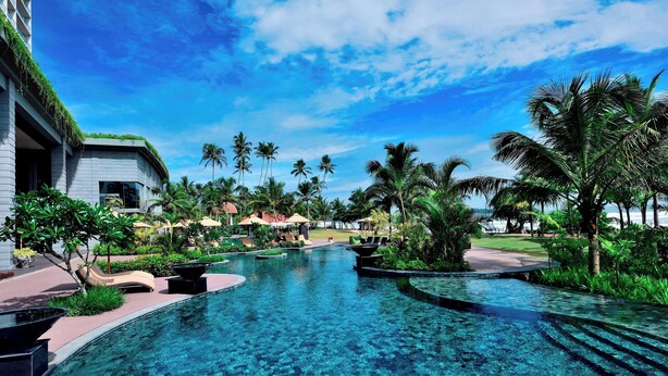 A clear pool with lounge chairs, palm trees, and a building with a green roof.