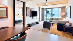 A modern hotel room with a dining area, a sofa set, a TV mounted on the wall, and a large window offering a view of the ocean.