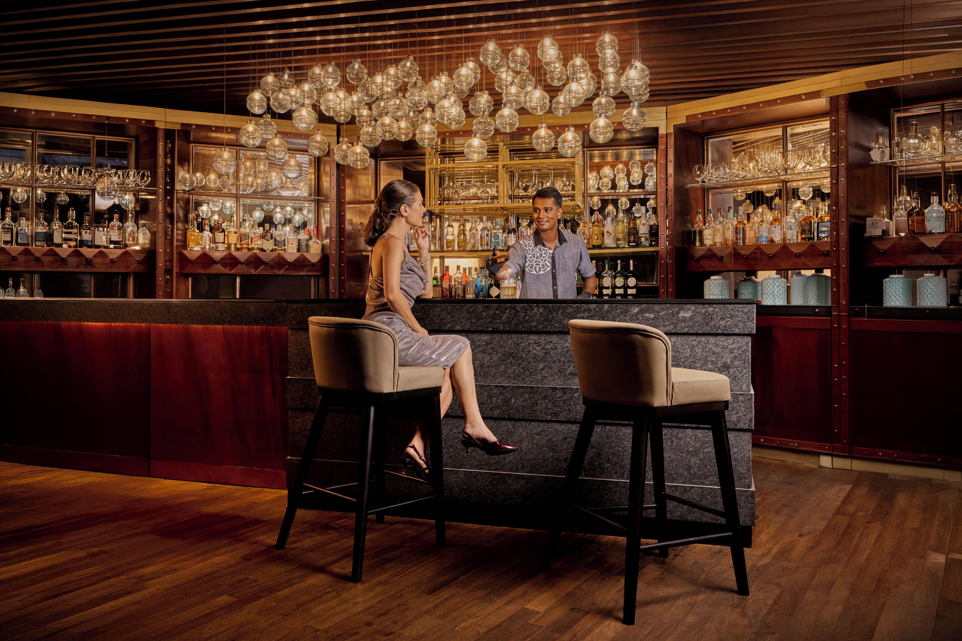 Een bar met twee hoge krukken, een barman en planken vol flessen en glazen.