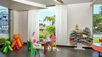 A child playing with toys at a table in a room with large windows, a tree-shaped bookshelf, and colorful chairs.