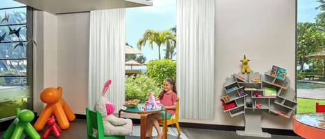 A child playing with toys at a table in a room with large windows, a tree-shaped bookshelf, and colorful chairs.