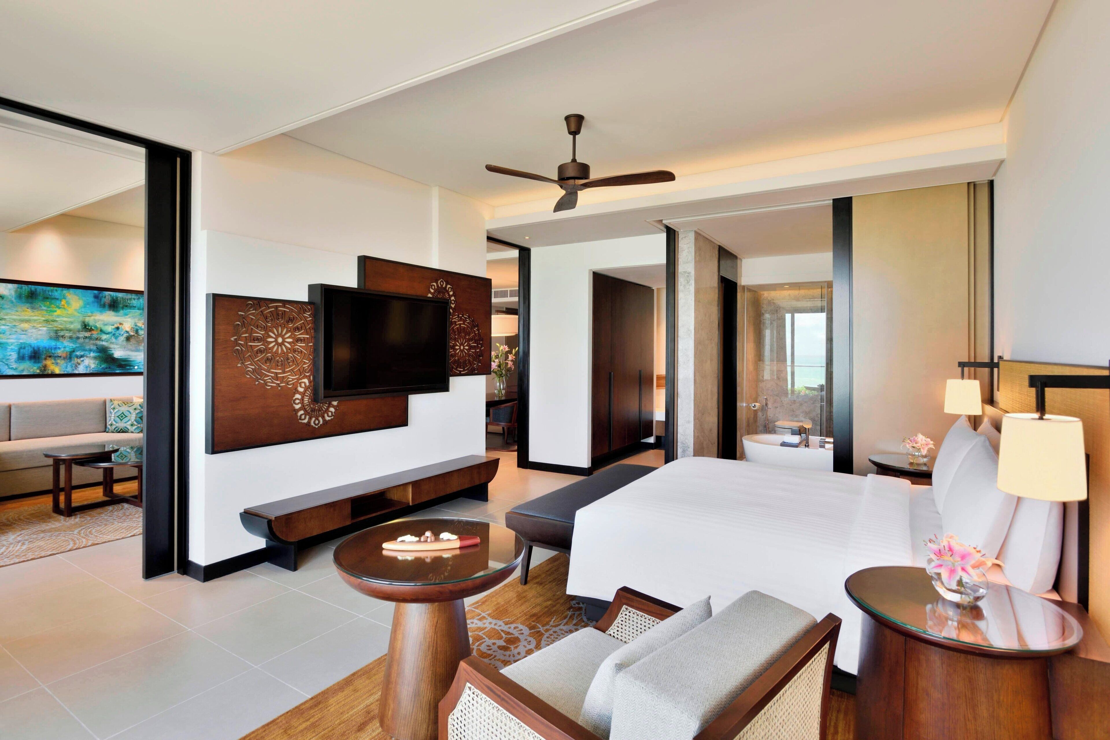 A modern hotel room with a large bed, a flat-screen TV mounted on the wall, a ceiling fan, and a seating area with a bench and a small table.
