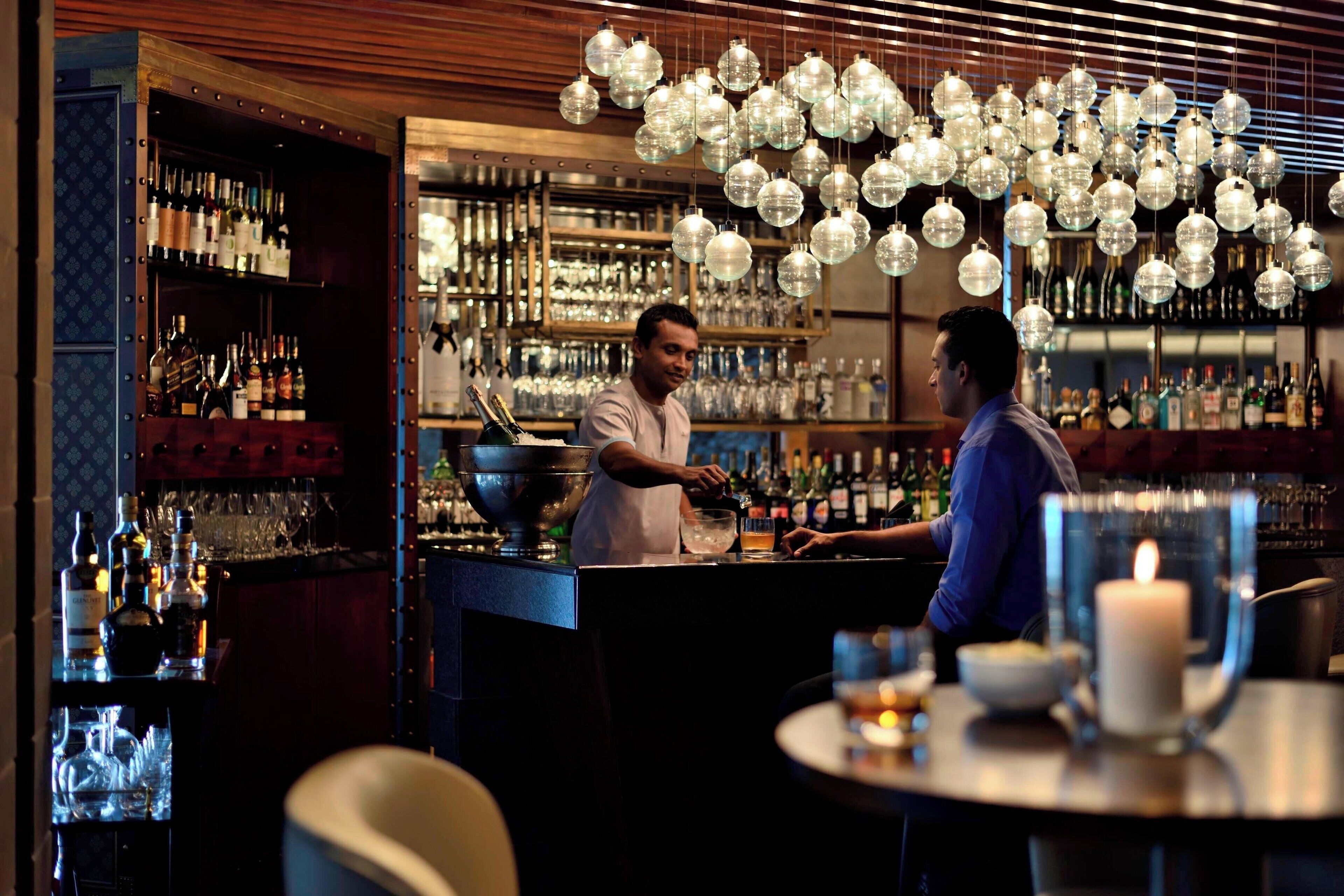 A bar with a bartender preparing drinks, shelves of bottles, and hanging light fixtures.