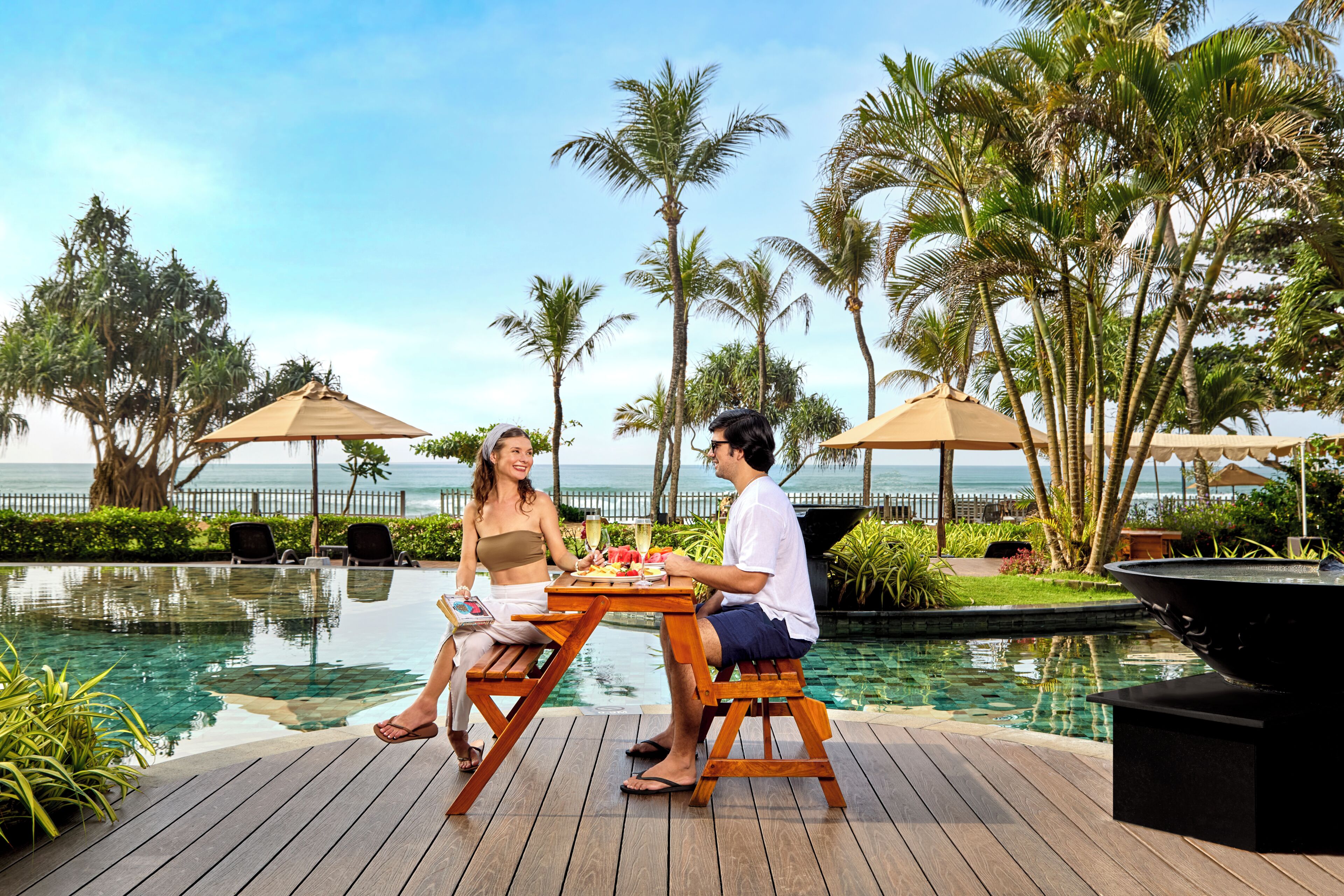 Un couple dînant au bord d’une piscine, avec vue sur l’océan, entouré de palmiers et de parasols.