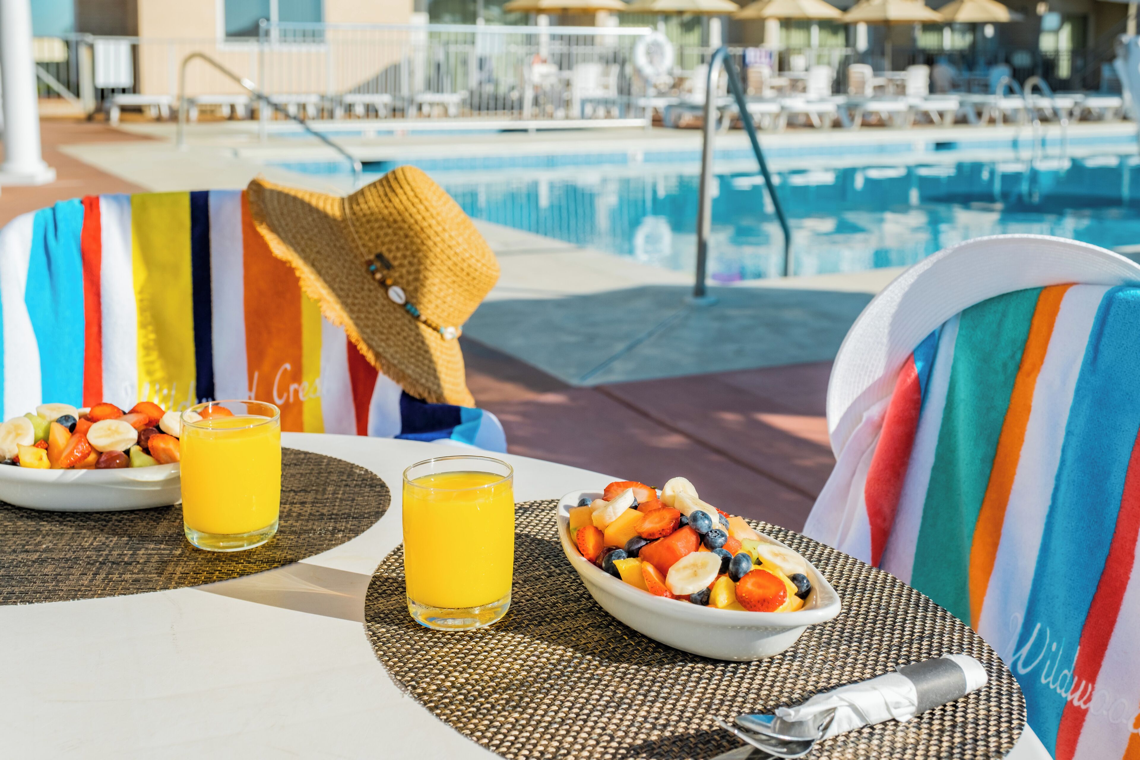 Ett matbord med en fruktskål och ett glas apelsinjuice, placerat nära ett område vid poolen med solstolar och parasoller.