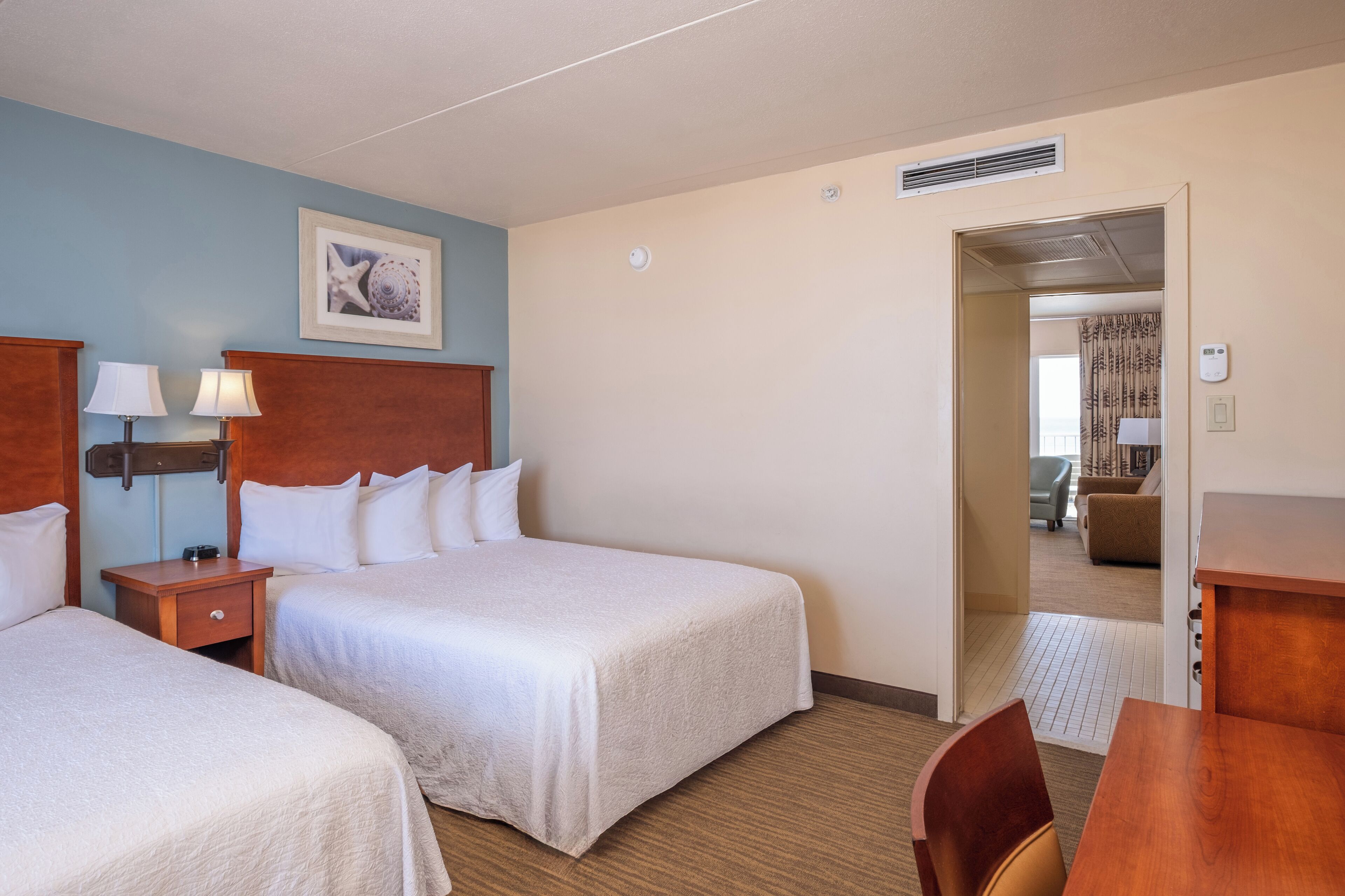Ein Hotelzimmer mit zwei Betten, einem Holzkopfstück, Nachttischlampen, einem gerahmten Bild an der Wand und Blick in den angrenzenden Wohnbereich.