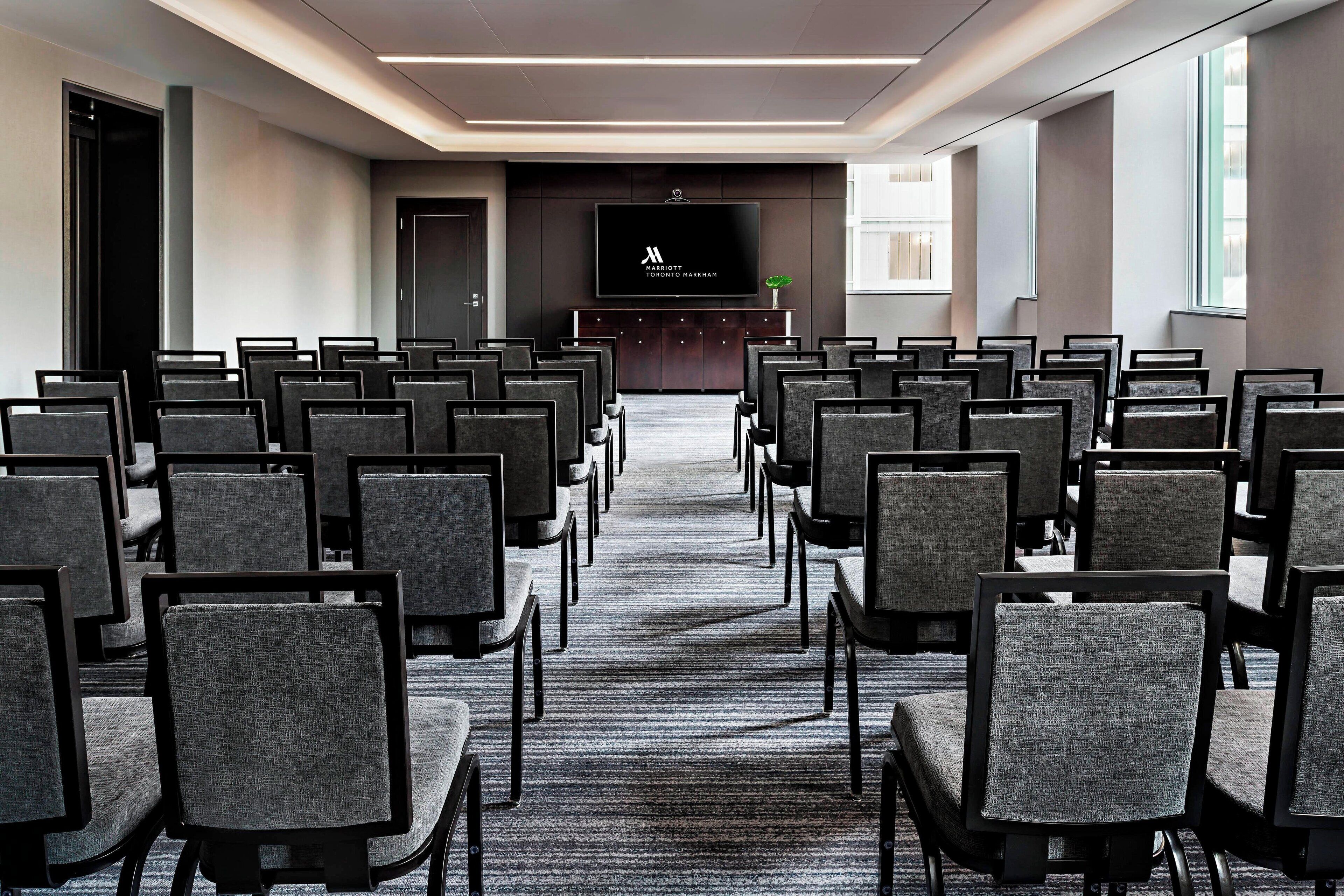 Sala de conferências com fileiras de cadeiras voltadas para uma tela, piso coberto de carpete e uma janela à direita.