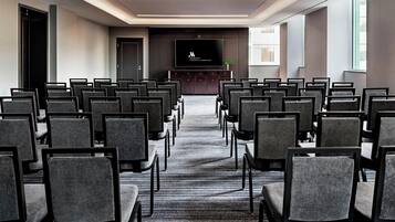 Sala de conferências com fileiras de cadeiras voltadas para uma tela, piso coberto de carpete e uma janela à direita.