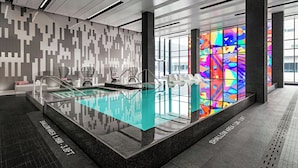 Un área de piscina interior moderna con diseño de pared geométrico, piscina de vidrio transparente y un ventanal de vidrio emplomado colorido.