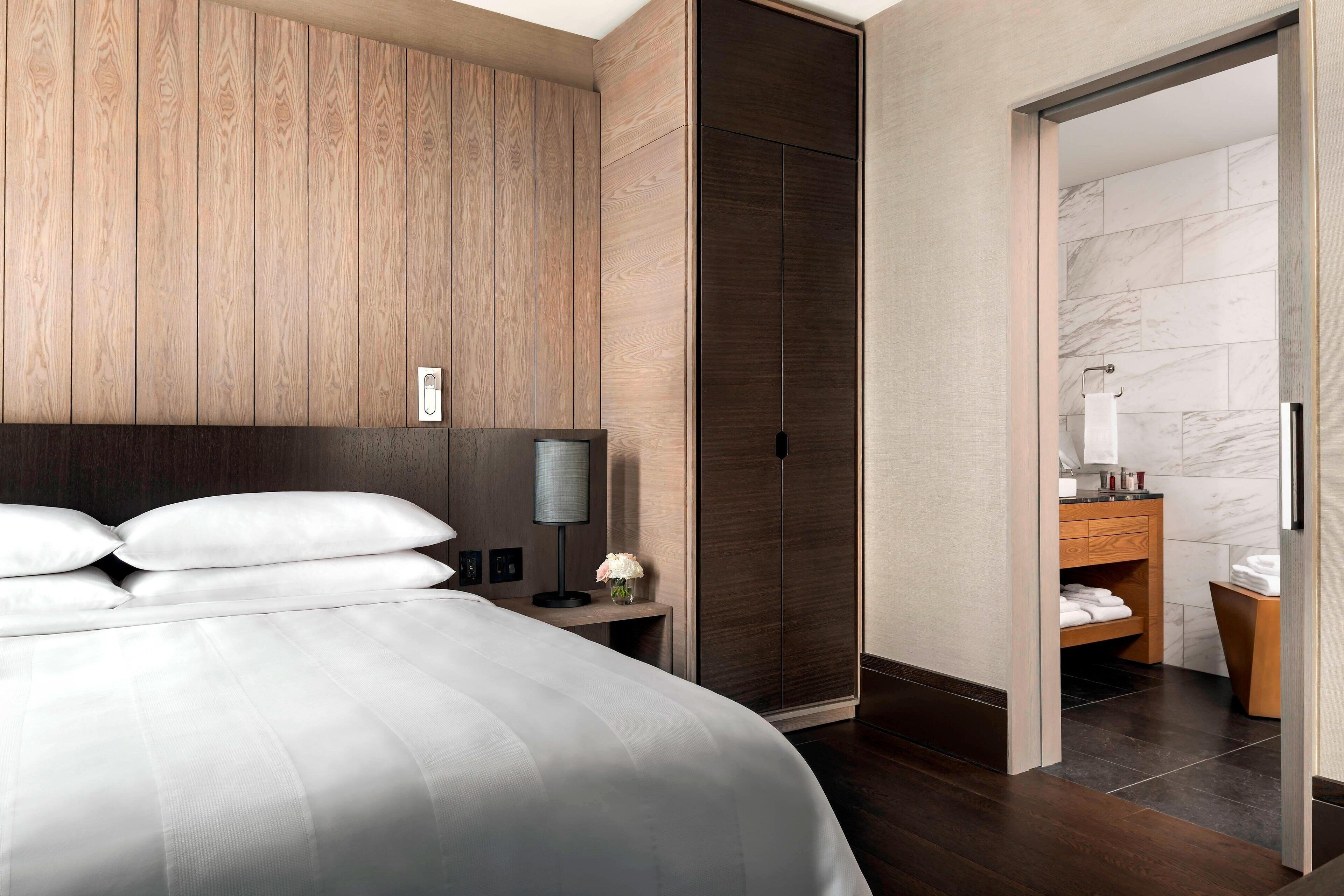 Et moderne hotelværelse med en stor seng, vægpaneler i træ, mørkt trægulv, et natbord med lampe og et badeværelse, der kan ses gennem en åben dør.