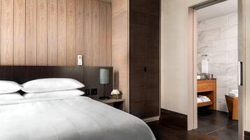 Et moderne hotelværelse med en stor seng, vægpaneler i træ, mørkt trægulv, et natbord med lampe og et badeværelse, der kan ses gennem en åben dør.
