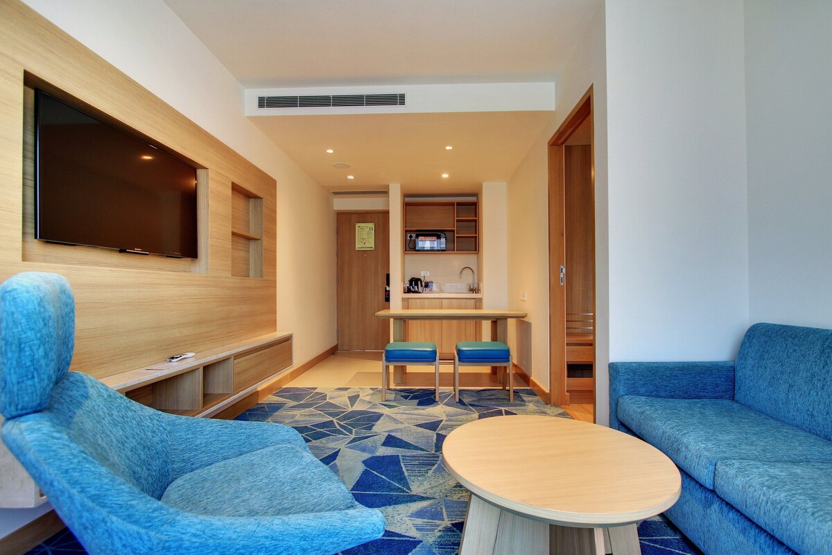 A modern living room with a flat-screen TV, blue upholstered chairs, a round coffee table, and a kitchen area in the background.