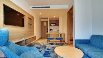 A modern living room with a flat-screen TV, blue upholstered chairs, a round coffee table, and a kitchen area in the background.