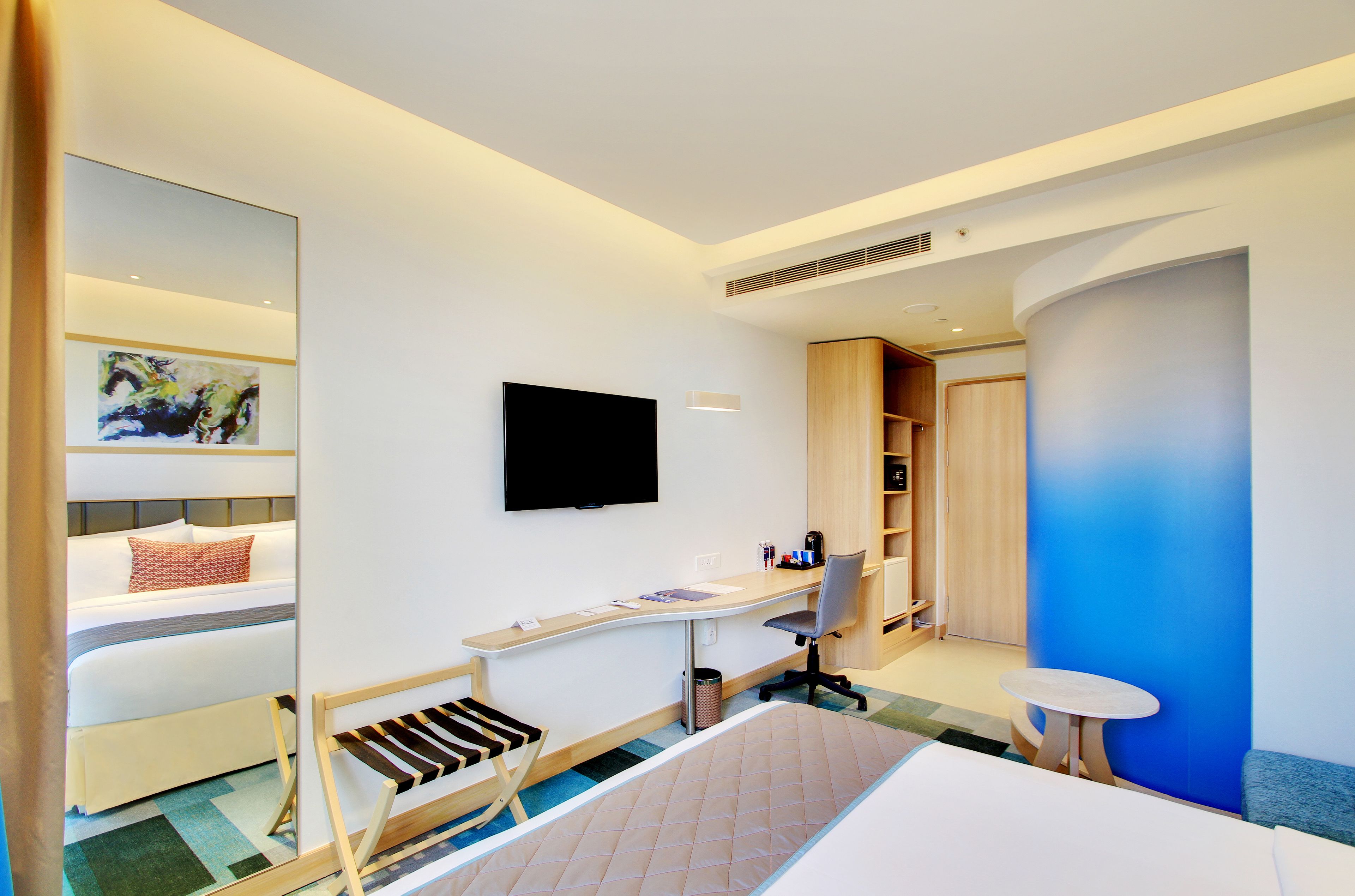 A hotel room with a bed, a desk, a chair, a TV, a small table, and a bookshelf.
