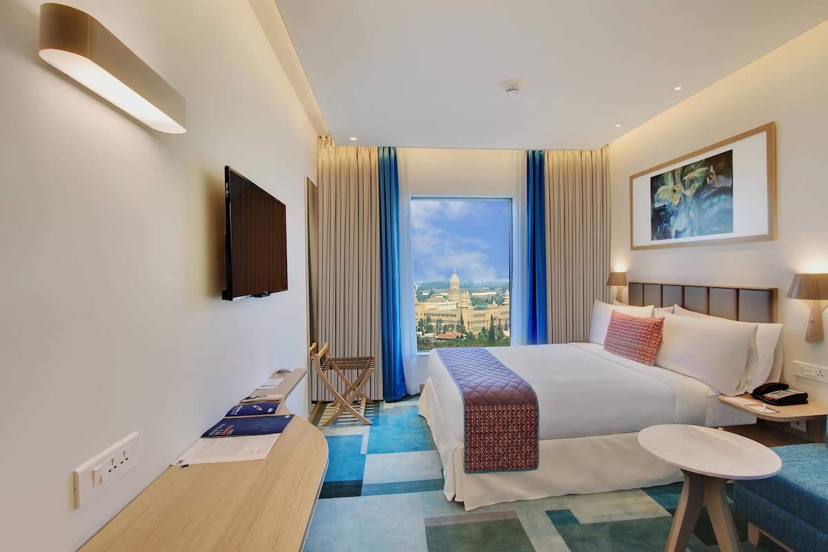 A hotel room with a large bed, a desk, a chair, a TV, and a view of a cityscape.