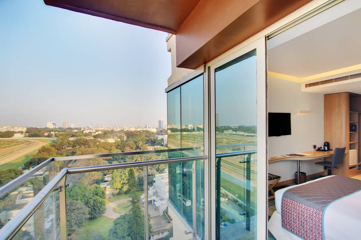 A modern hotel room with a balcony, large windows, a desk, and a bed.
