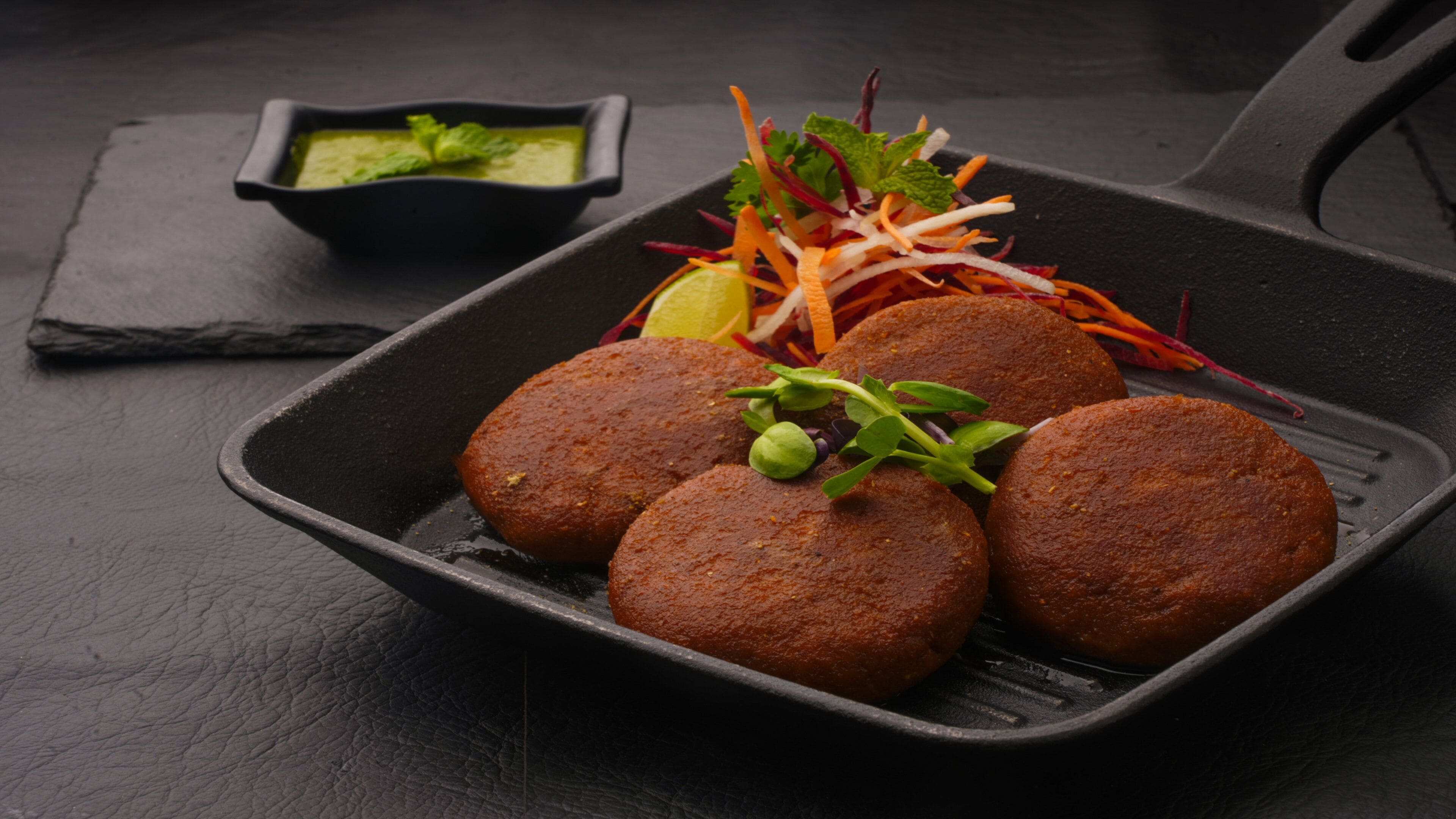 A black skillet with four round, brown patties, a small bowl of green sauce, and a side salad with shredded carrots and greens.