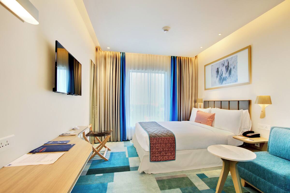 A hotel room with a bed, a desk, a chair, a TV, and a window with curtains.