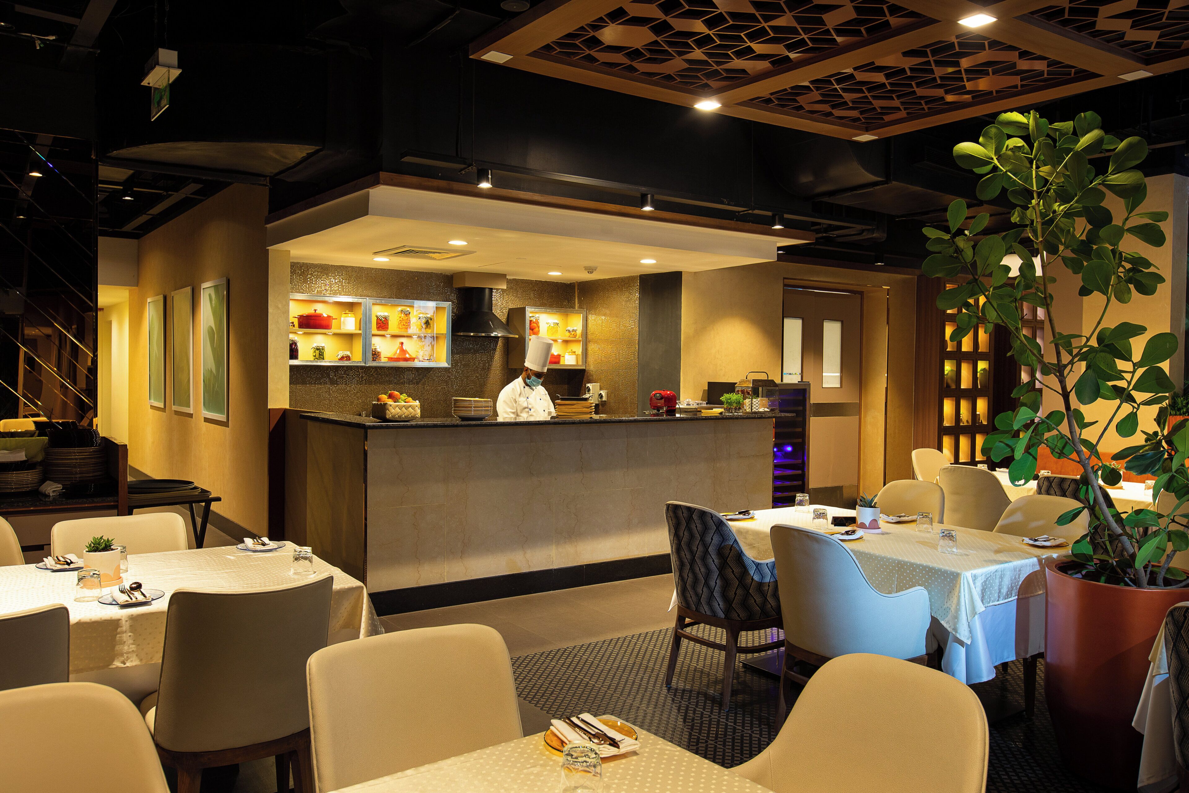 A restaurant interior with a chef at the counter, tables with chairs, and a potted plant.