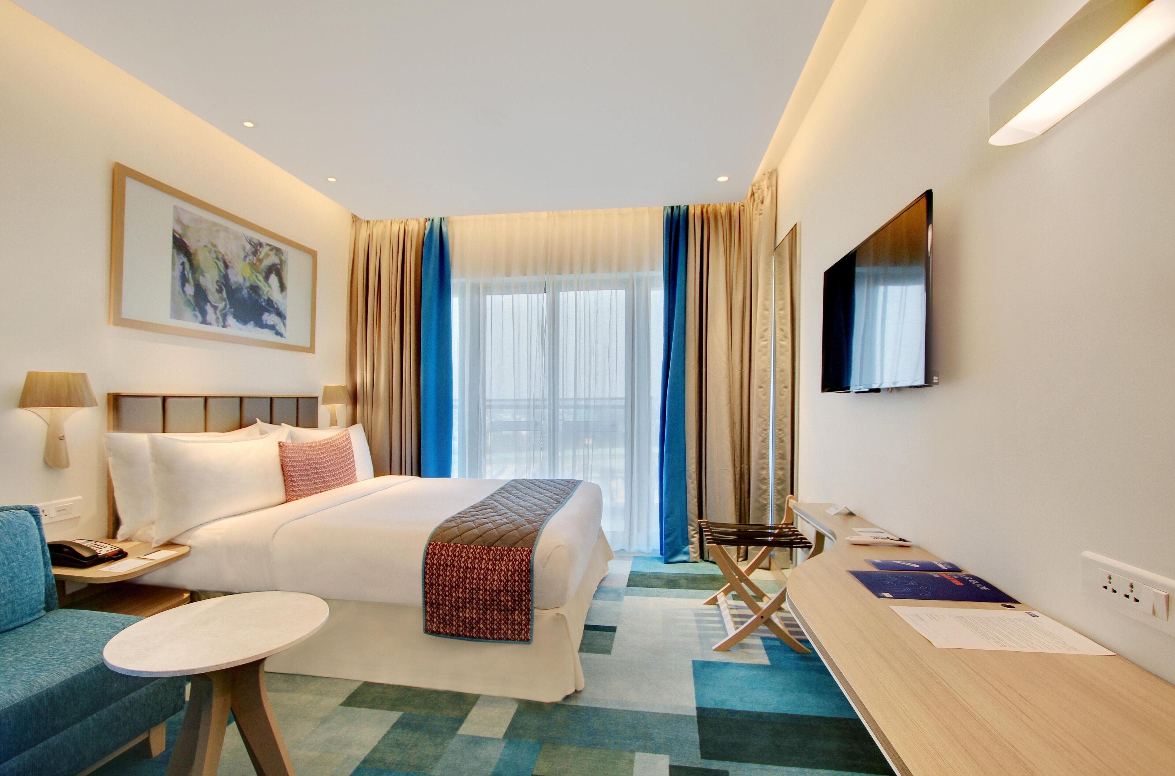 A hotel room with a bed, bedside table, lamp, chair, TV, and a painting on the wall.