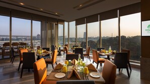 Un área de comedor con mesas dispuestas para el desayuno, amplias ventanas con vistas a la ciudad y un cartel que indica la ubicación como Holiday Inn Bengaluru Airport.