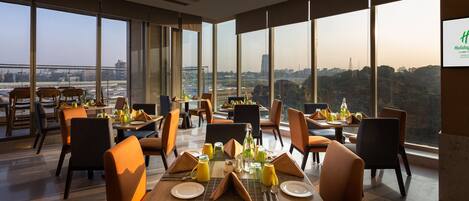 Un área de comedor con mesas dispuestas para el desayuno, amplias ventanas con vistas a la ciudad y un cartel que indica la ubicación como Holiday Inn Bengaluru Airport.