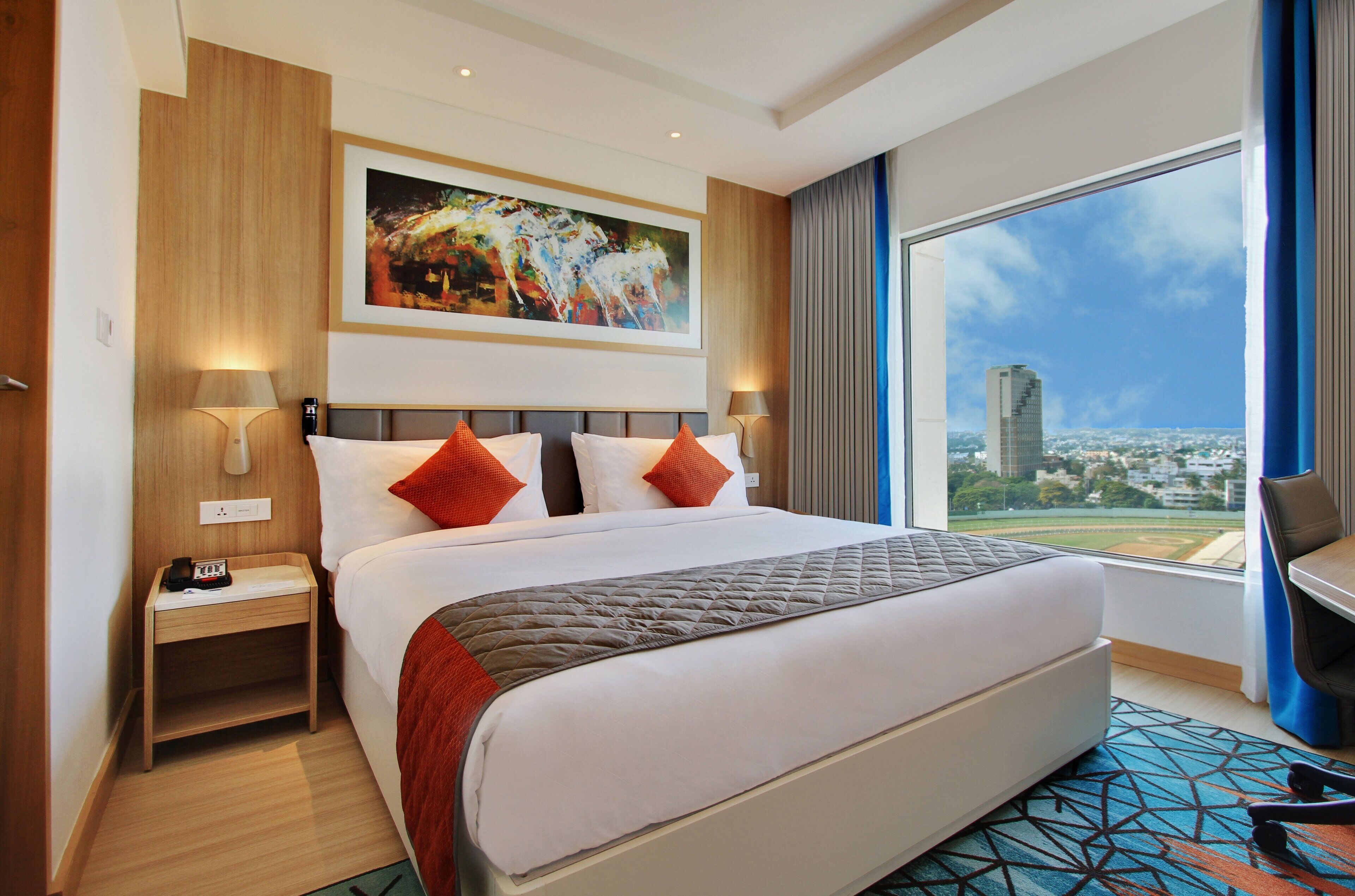 A hotel room with a large bed, a nightstand, a desk, a chair, and a large window offering a city view.