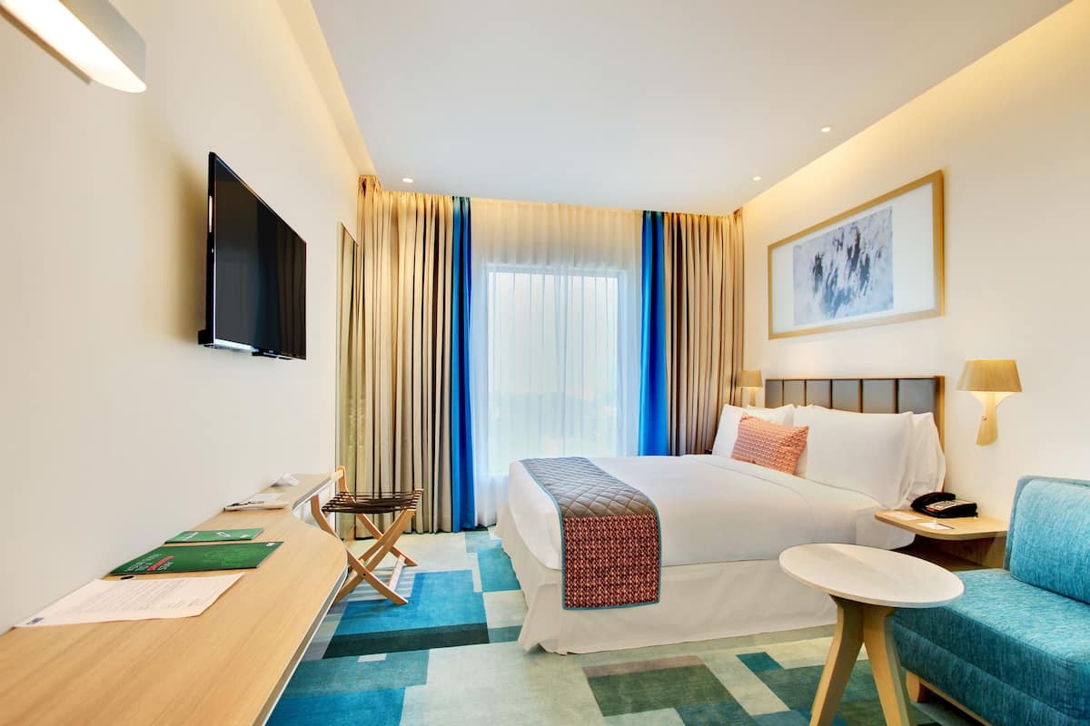 A hotel room with a bed, a desk, a TV, a chair, a sofa, and a window with curtains.