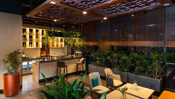 Un área de bar moderna con asientos, una cocina al fondo y ventanas amplias con persianas.