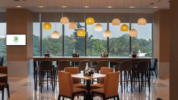 A hotel dining area with round tables and chairs, large windows with a view of greenery, and pendant lighting.