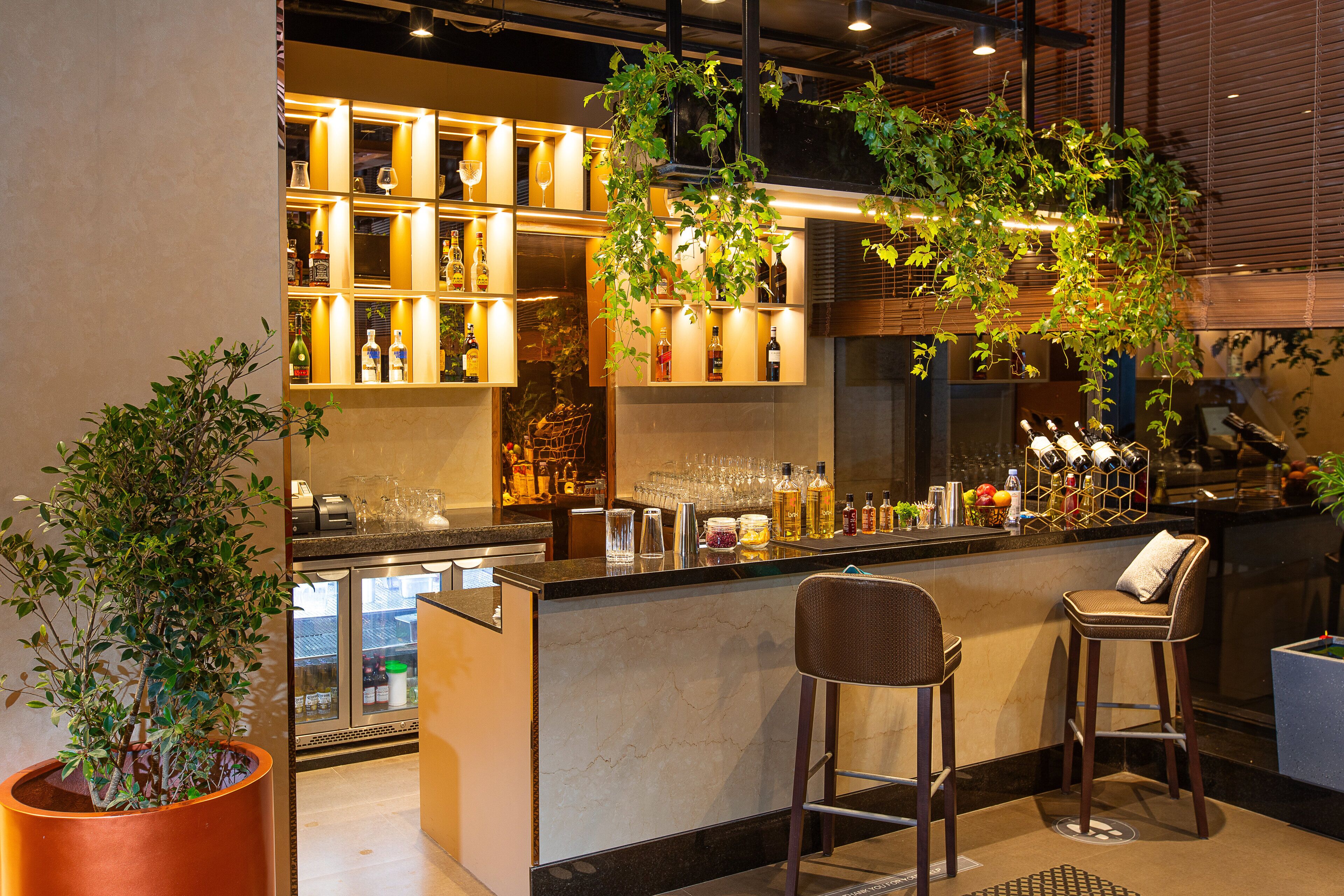 Eine Bar mit Marmorplatte, zwei Barhocker, Regalen mit Flaschen und Gläsern sowie einer großen Topfpflanze.