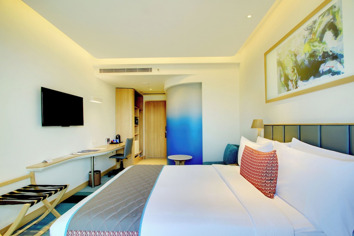 A hotel room with a bed, a desk, a chair, a TV, and a painting on the wall.