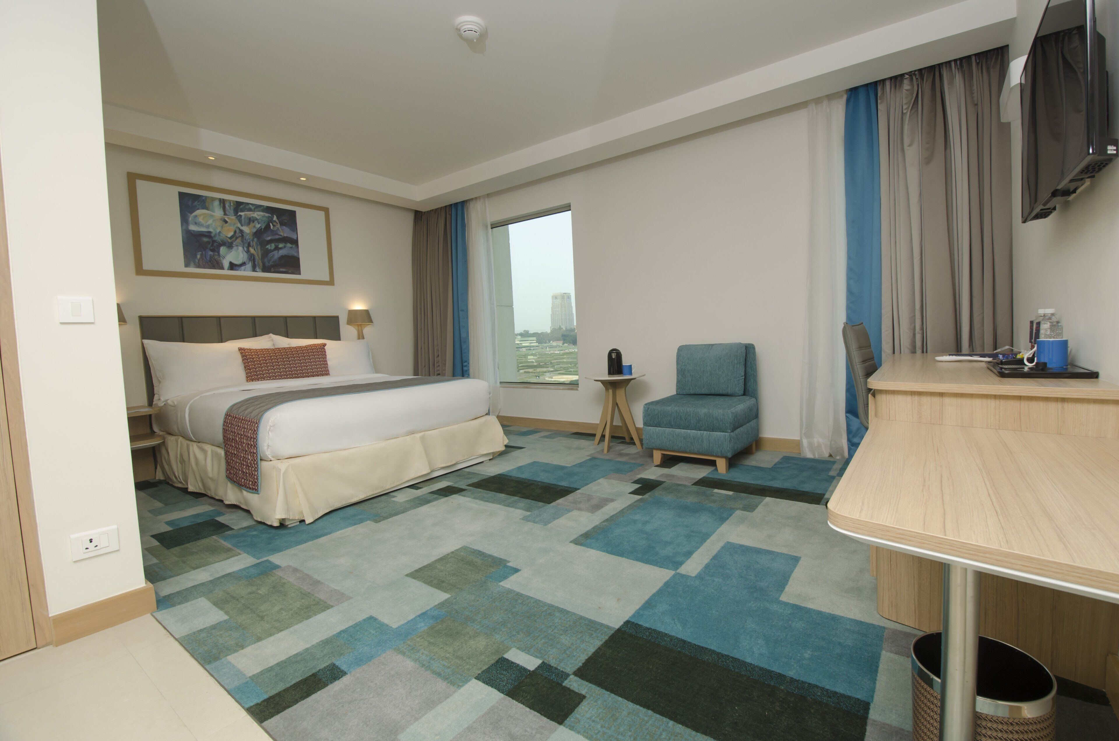 A hotel room with a bed, a desk, a chair, a TV, and a window with curtains.