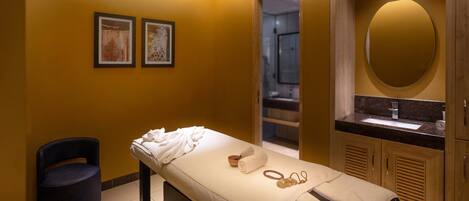 Un cuarto de spa con una camilla de masaje, una silla azul, un lavabo y obras de arte enmarcadas en la pared.