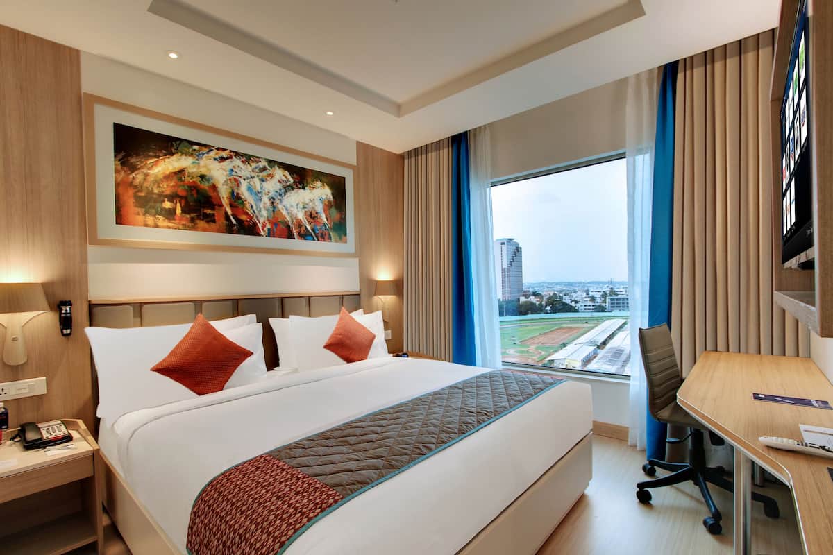 A hotel room with a large bed, a desk, a chair, a painting on the wall, and a view of a cityscape through the window.