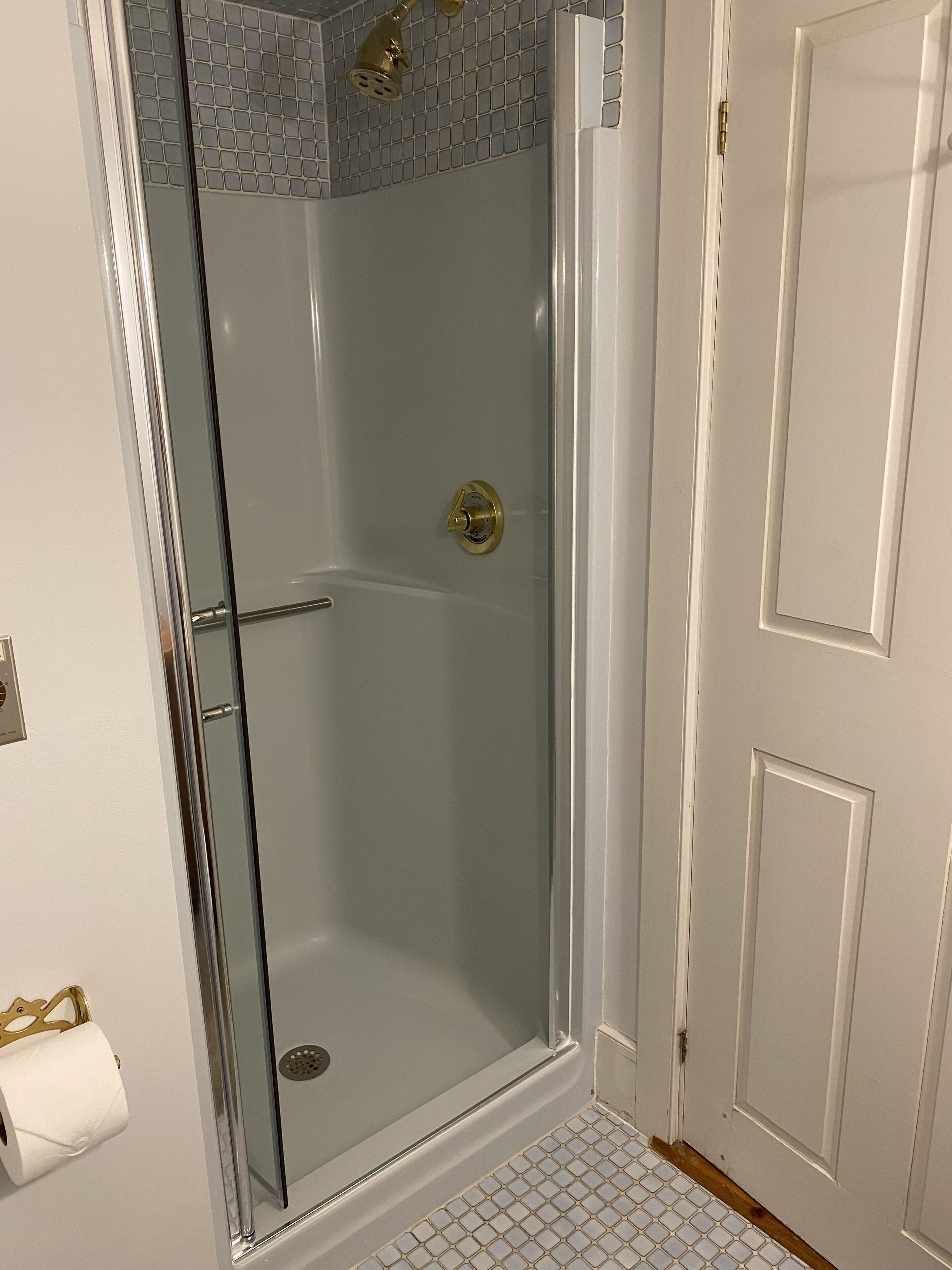 Una ducha con paredes de vidrio, un cabezal de ducha dorado, una puerta blanca con pomo dorado y un piso de baldosas blancas.