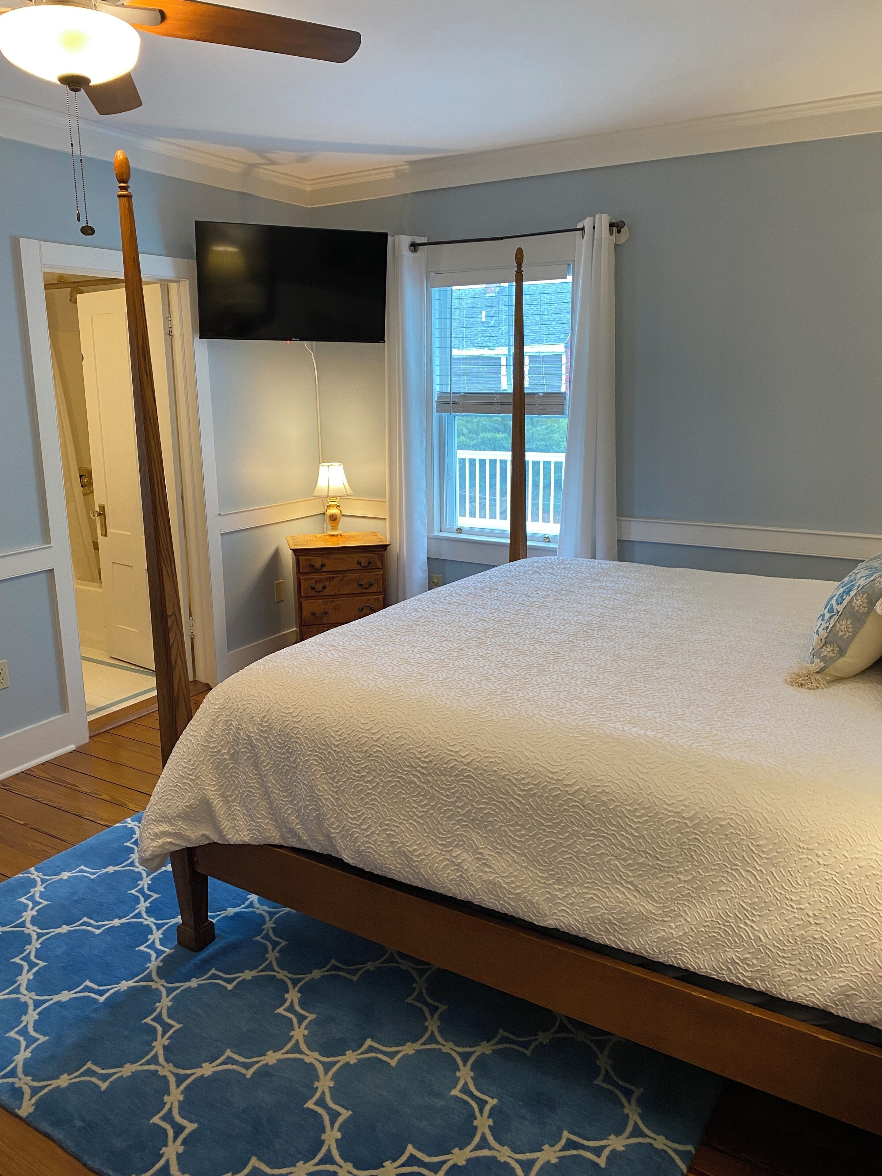 A bedroom with a bed, a nightstand, a lamp, a ceiling fan, a TV, and a window with a view of a balcony.